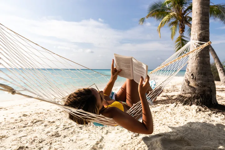 Egy bikini-s nő könyvet olvas a pálmafák közé kifeszített függőágyban egy fehér homokos, trópusi tengerparton.