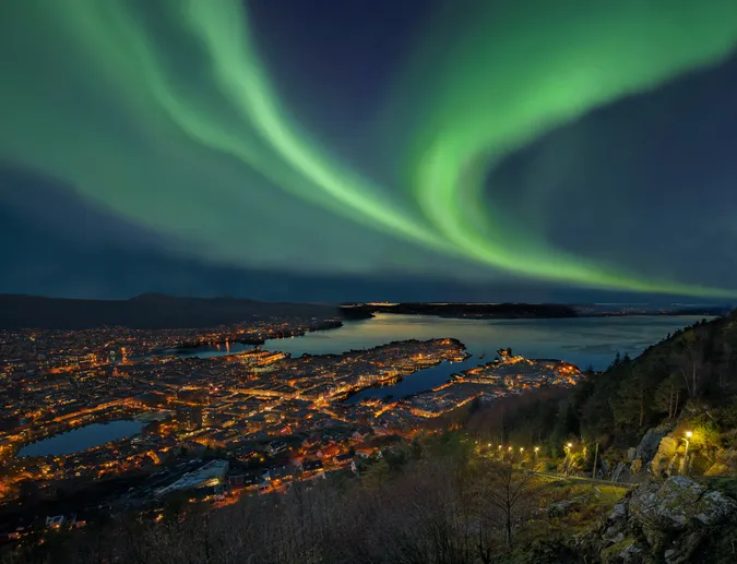 A zöldellő sarki fény örvénylik Bergen kivilágított éjszakai városa, a fjordok és a környező hegyek felett a Fløyen-hegyről nézve.