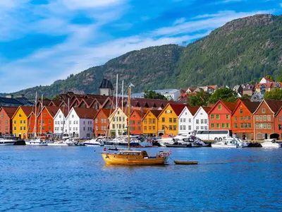 Bergen színes Bryggen negyedének faházai a vízparton, előtérben egy fahajóval, háttérben pedig zöldellő hegyoldallal.