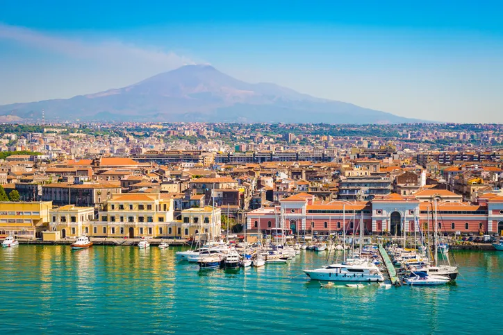 Catania kikötője jachtokkal és színes épületekkel Szicíliában, a háttérben a füstölgő Etna vulkánnal.