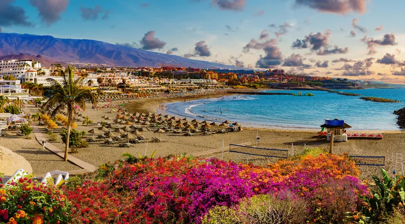 A tenerifei Costa Adeje tengerpartja színes virágokkal, napernyőkkel és a háttérben magasodó hegyekkel.