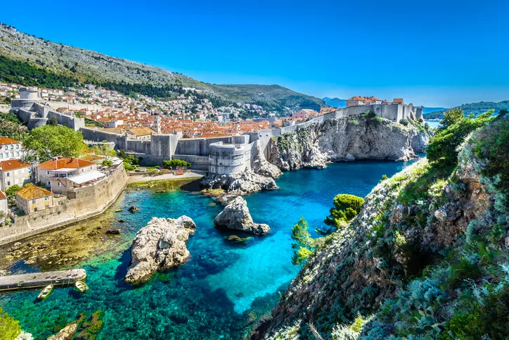 Dubrovnik monumentális várfalai és a sziklás, kristálytiszta vizű öböl látképe a zöldellő partoldalról, ragyogó napsütésben.
