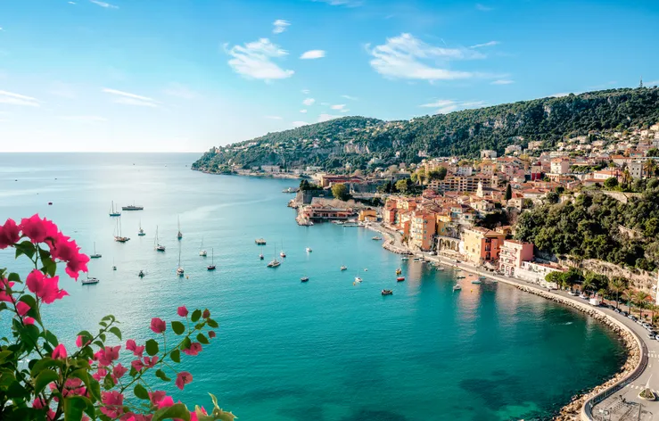 Panorámás kilátás Villefranche-sur-Mer színes házaira és a Francia Riviéra azúrkék öblére, előtérben pink virágokkal.