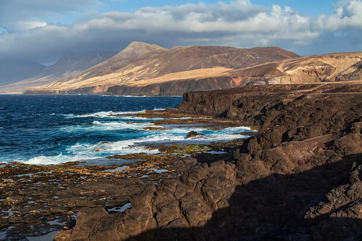 Fuerteventura sziklás, vulkanikus tengerpartja a Cofete strand közelében, háttérben a felhőkbe burkolózó Jandía-hegységgel.