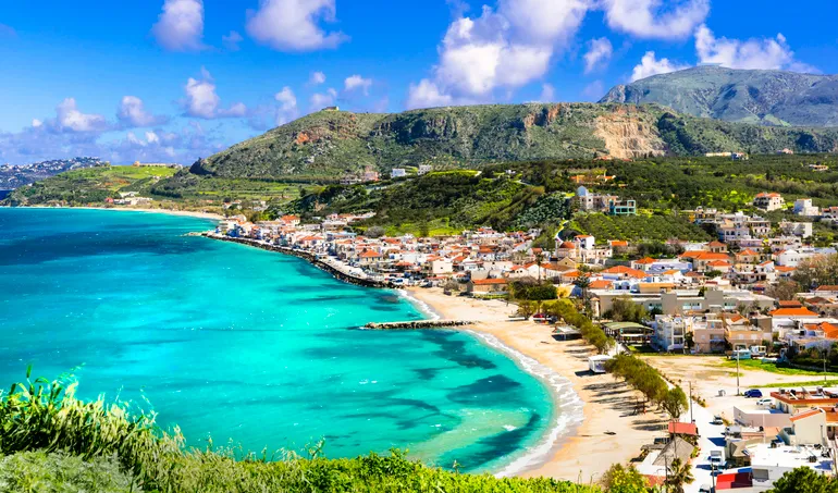 Panorámás kilátás a krétai Kalyves tengerpartjára, türkizkék vízzel és zöld hegyekkel a háttérben.