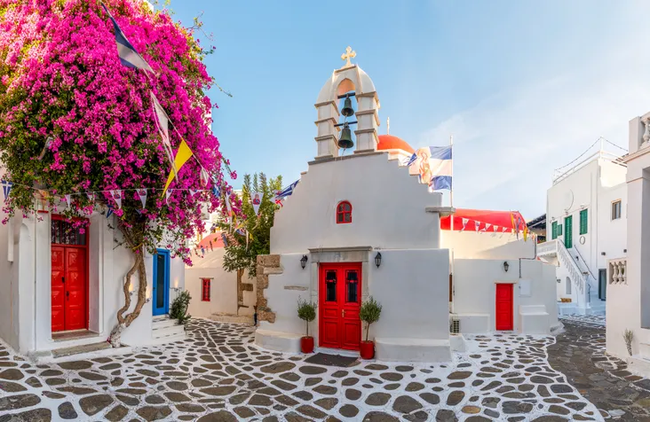 Görög ortodox templom fehér falaival, harangtornyával és piros ajtójával Mikonoszon, mellette vibráló pink murvafürttel (bougainvillea).
