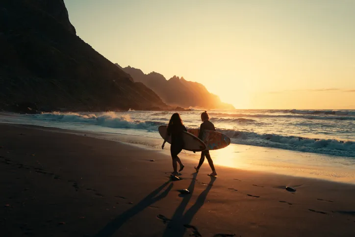 Két szörfös sétál a tenerifei Playa de Benijo fekete homokos partján a naplemente fényében, sziklás háttérrel.