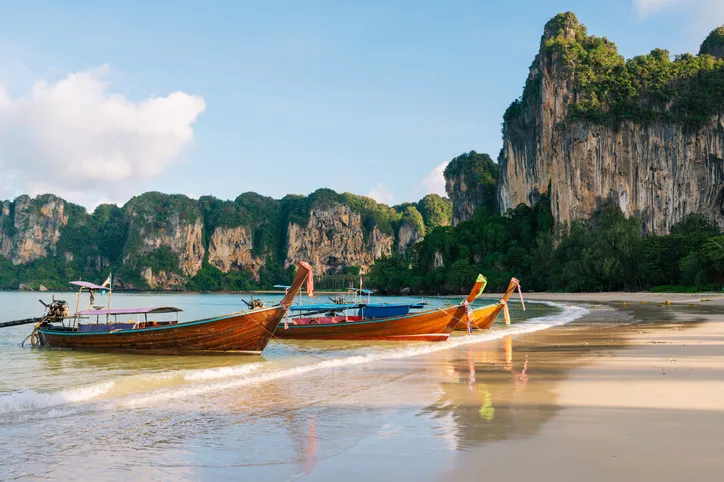 Hagyományos fából készült thaiföldi hosszúfarkú hajók (longtail boat) a Railay Beach homokos partján, háttérben sziklákkal.
